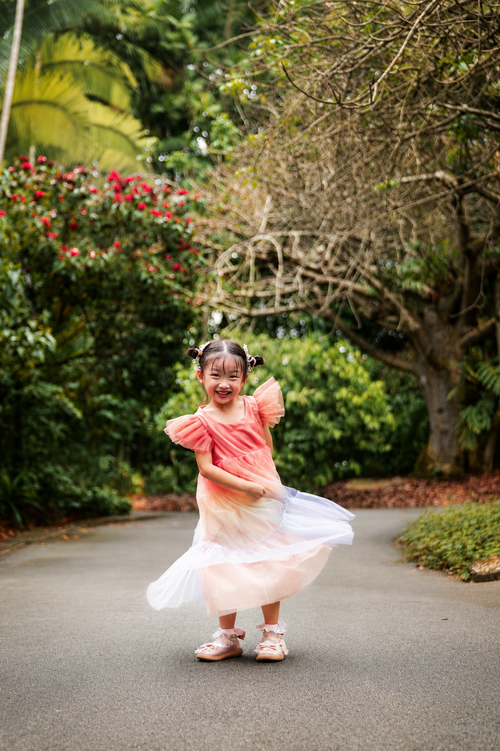 Happy little girl in pink gradient tulle dress twirling on a scenic garden pathway with lush trees.