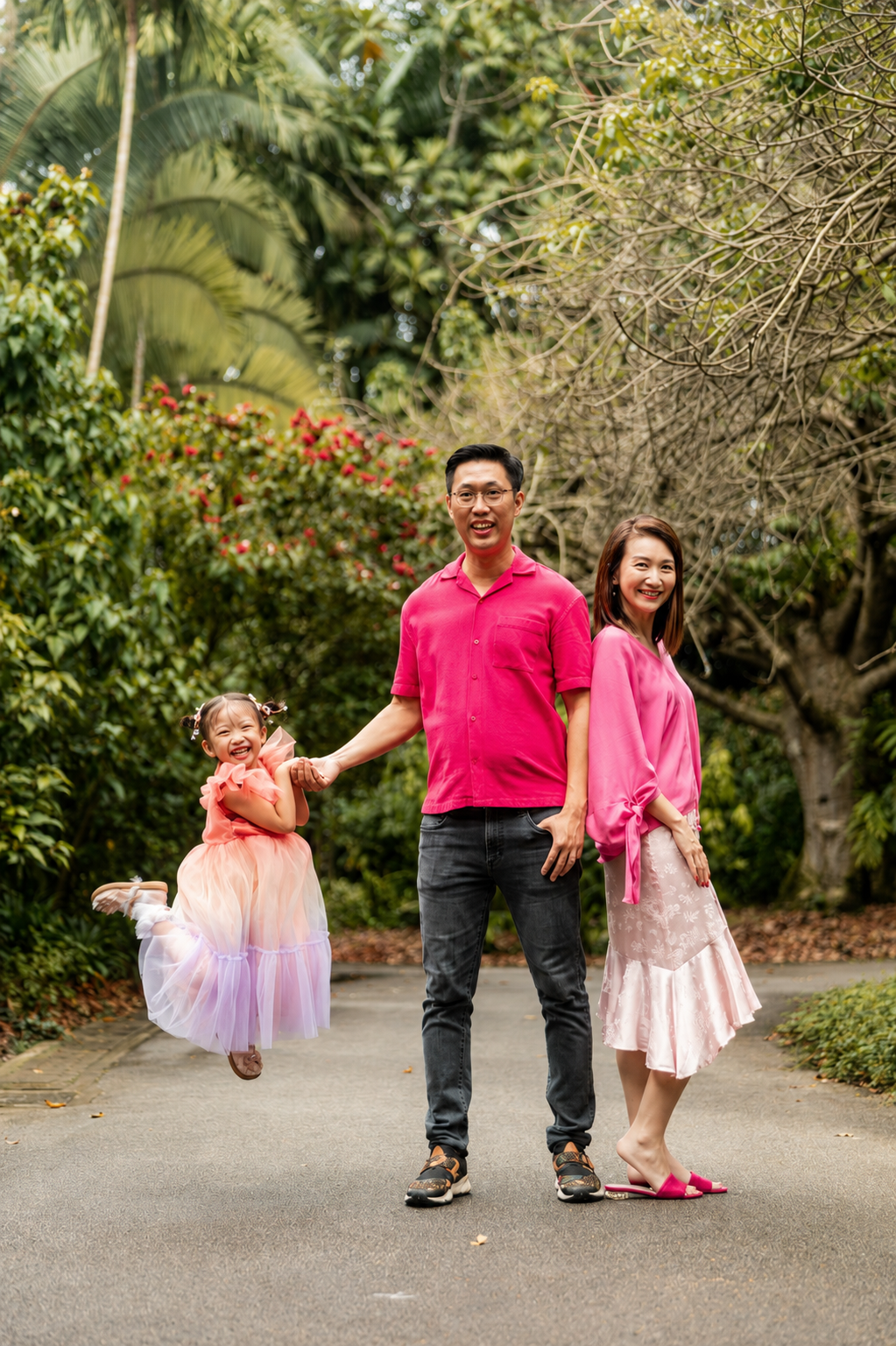 Happy family in pink outfits posing on a tree-lined park pathway with father swinging daughter.