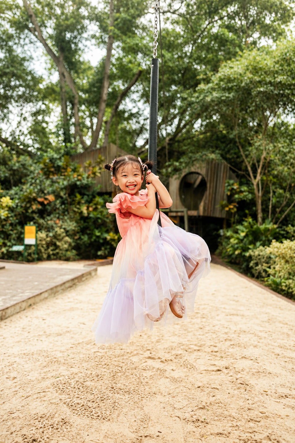 Smiling young girl in pink tulle dress enjoying a swing at a sandy playground surrounded by trees.