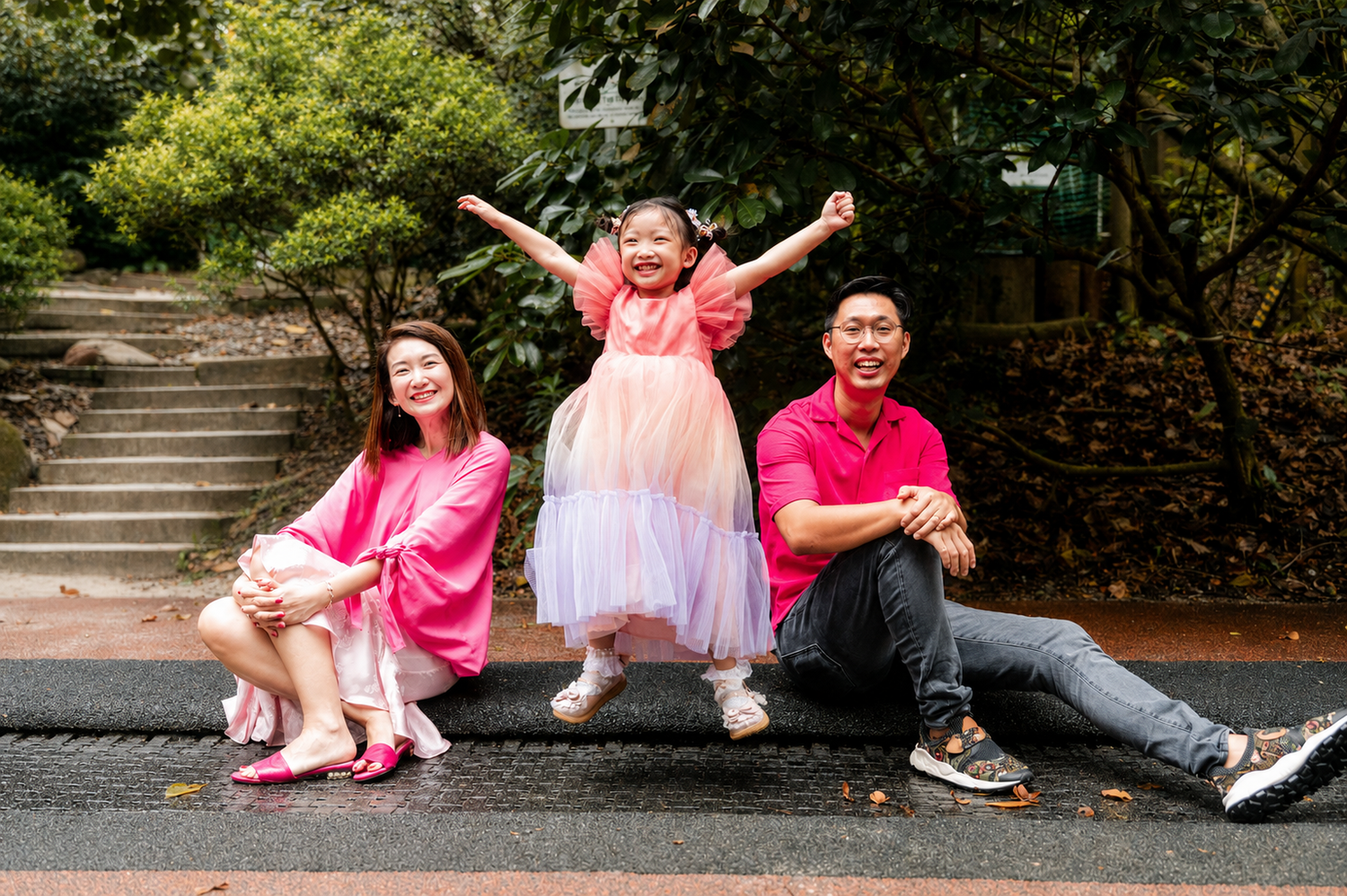 Joyful family of three posing playfully outdoors with daughter jumping between seated parents.