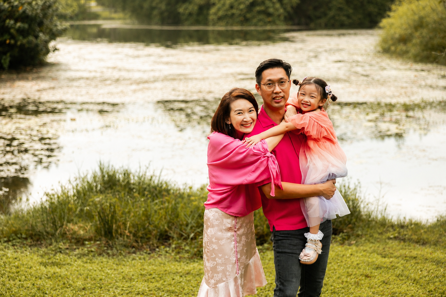 Happy Asian family of three posing by a serene pond, parents smiling with young daughter in pink dress.