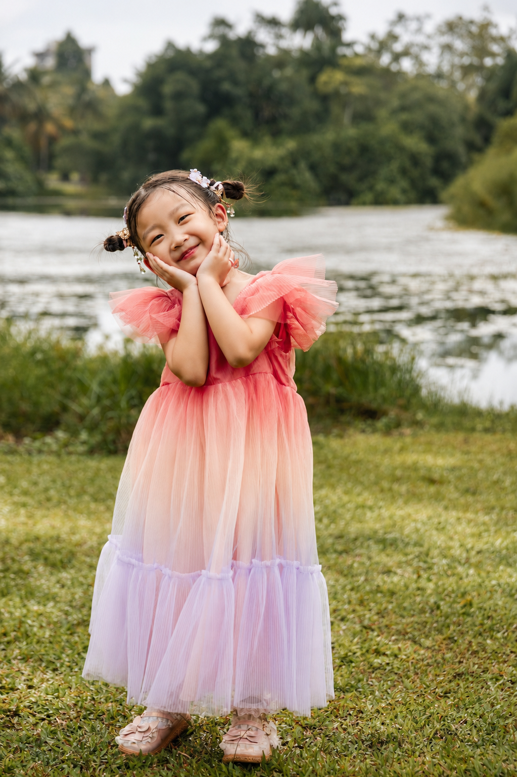 Adorable little girl in pink ombre tulle dress posing joyfully by a peaceful lake in a garden.