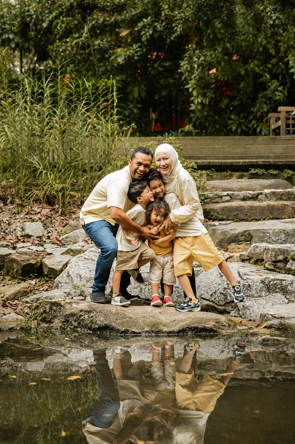 Loving multiracial family of four hugging on stone steps by a reflective pond in lush greenery.