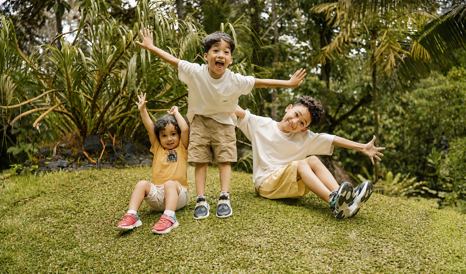 Three joyful children playing on green grass with arms outstretched in a tropical garden setting.