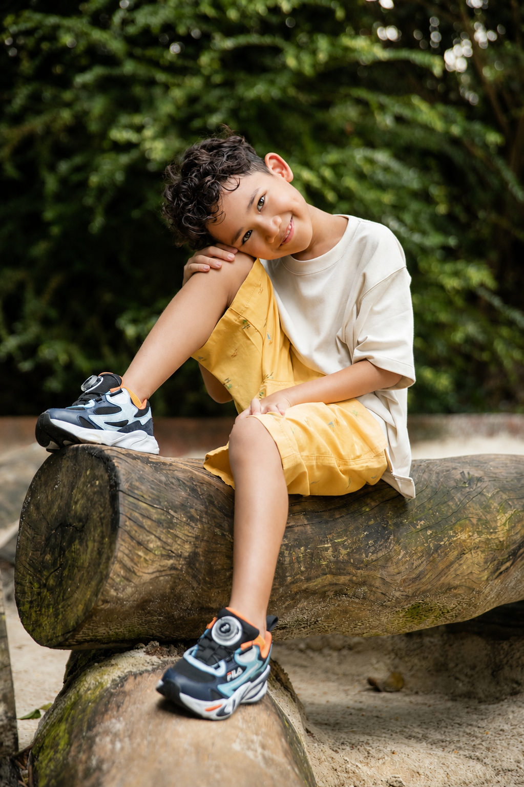 A young boy with curly hair sits on a log outdoors, wearing yellow shorts and sporty sneakers, smiling at the camera.