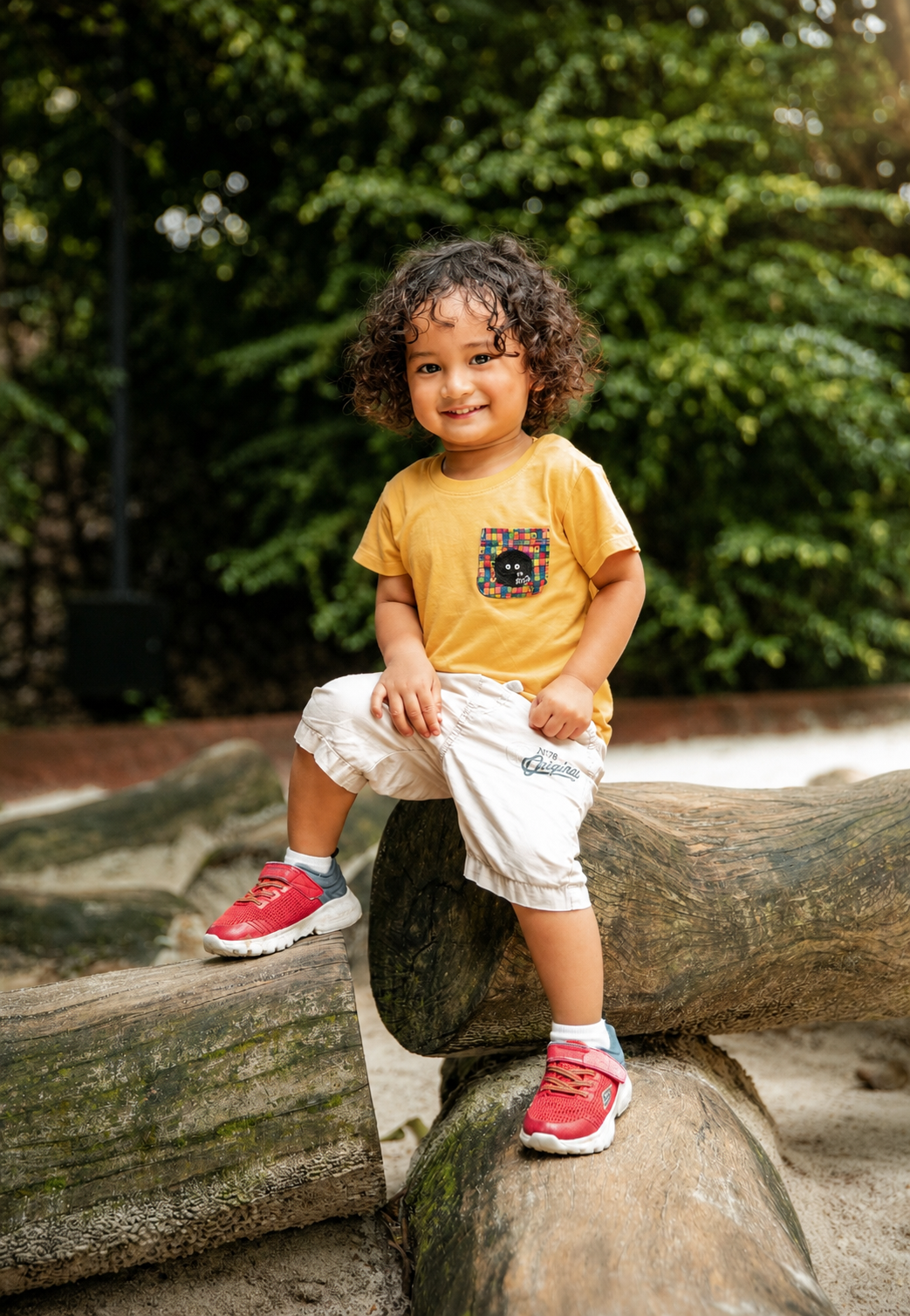 A toddler with curly hair sits on a log wearing a yellow graphic tee, white pants, and red sneakers in a park setting.