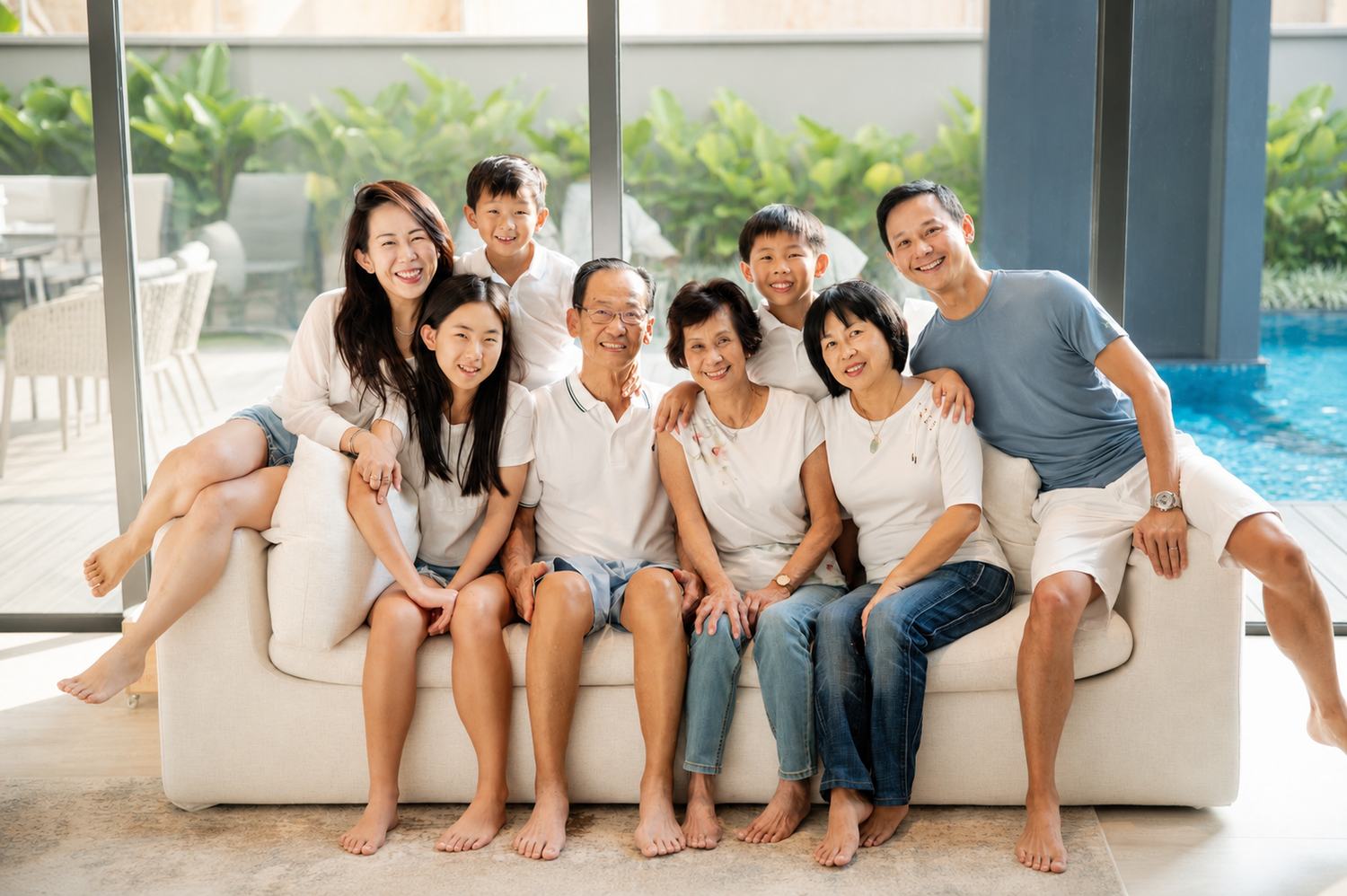 A large Asian family of nine smiles together on a white sofa near an indoor pool in a bright home.