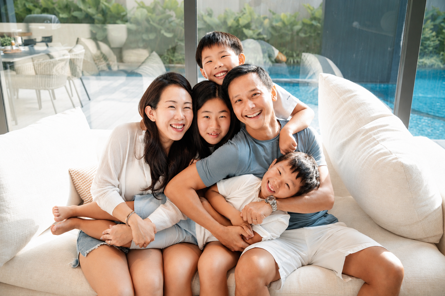 A cheerful Asian family of five cuddles closely together on a white sofa, laughing and smiling near an indoor pool.