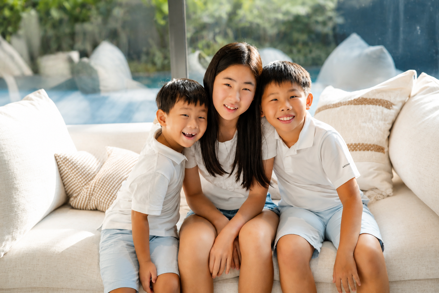 Three Asian siblings smile and hug each other while sitting together on a white sofa in a bright living room.