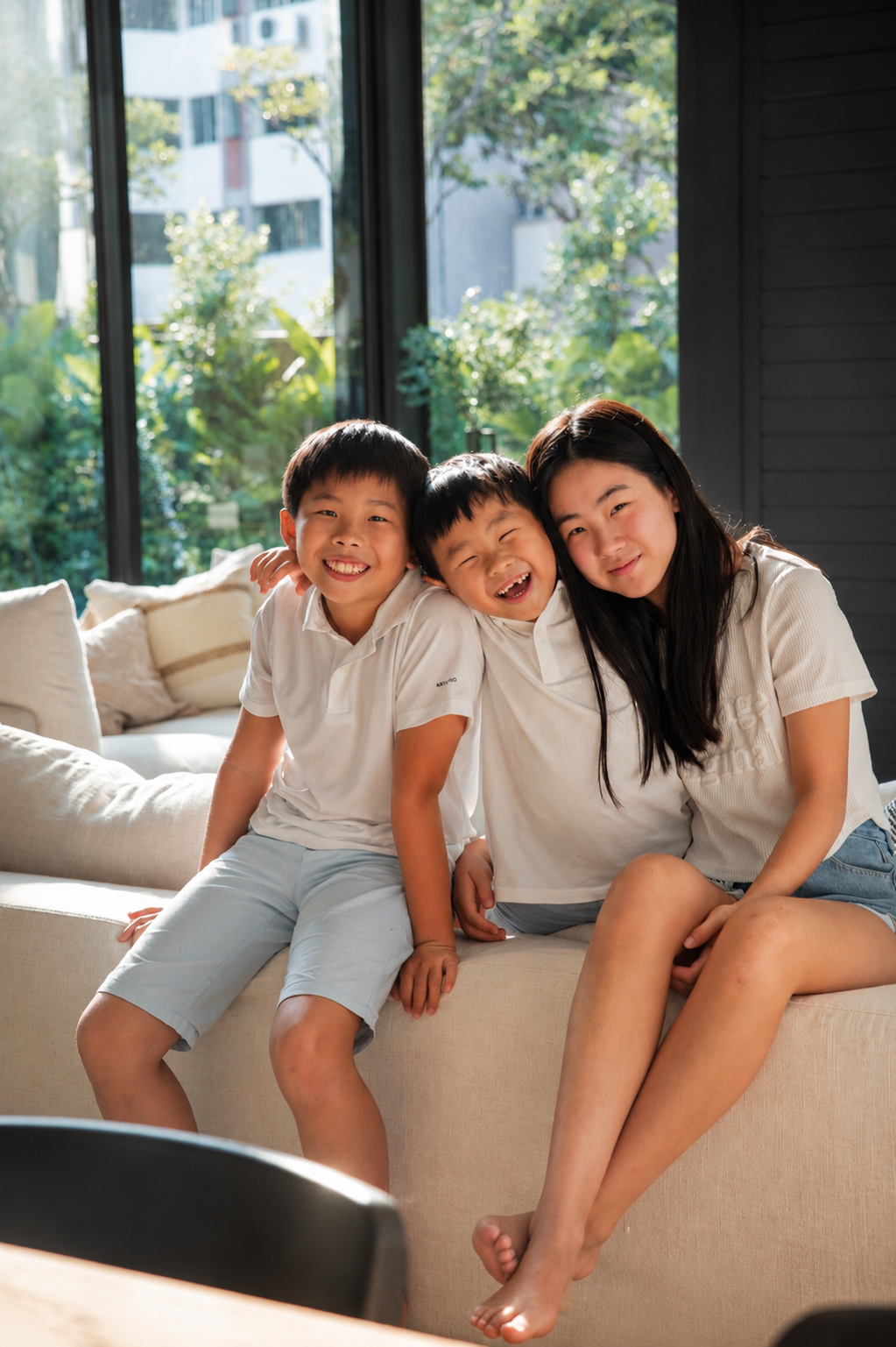 Two boys and a teenage girl laugh and pose together on a white sofa in a sunlit modern living room.