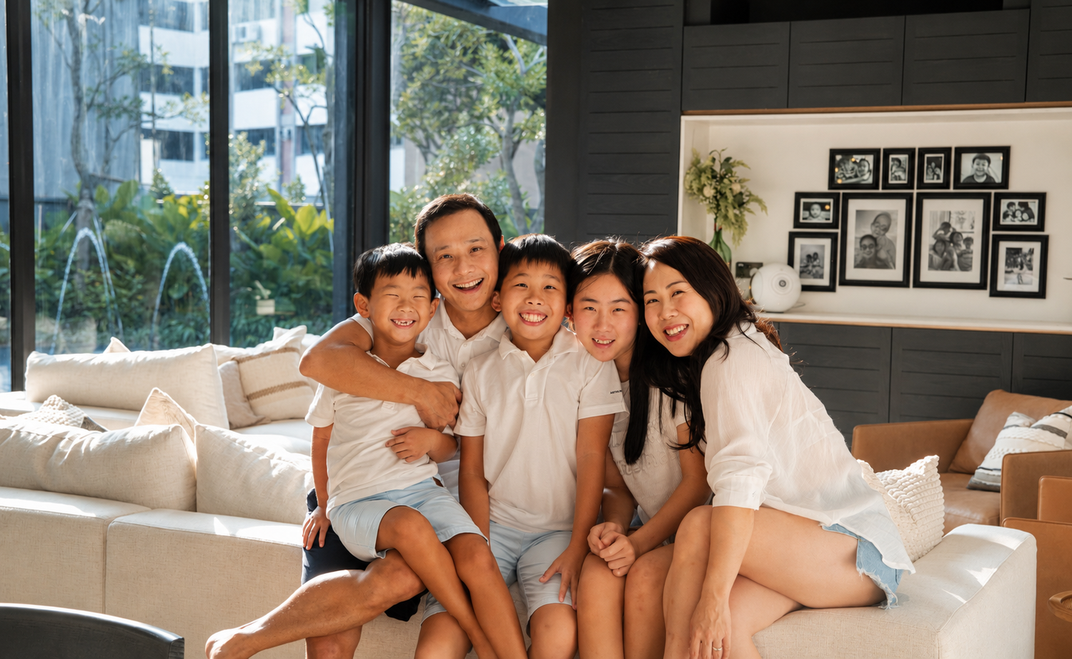 A smiling Asian family of four hugs together on a white sofa in a stylish modern living room.
