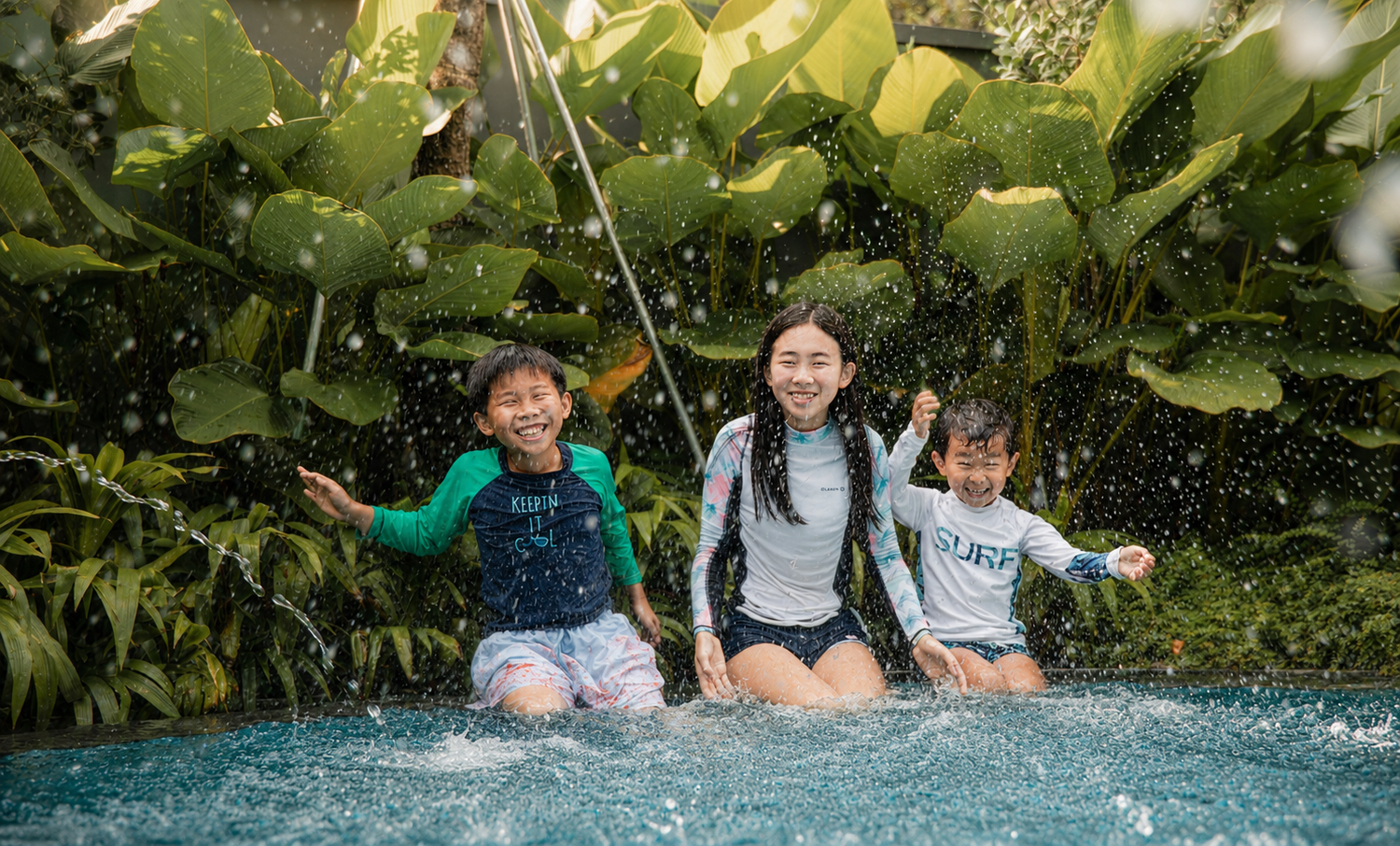 Joyful woman and two children splashing and laughing in a lush tropical outdoor pool.