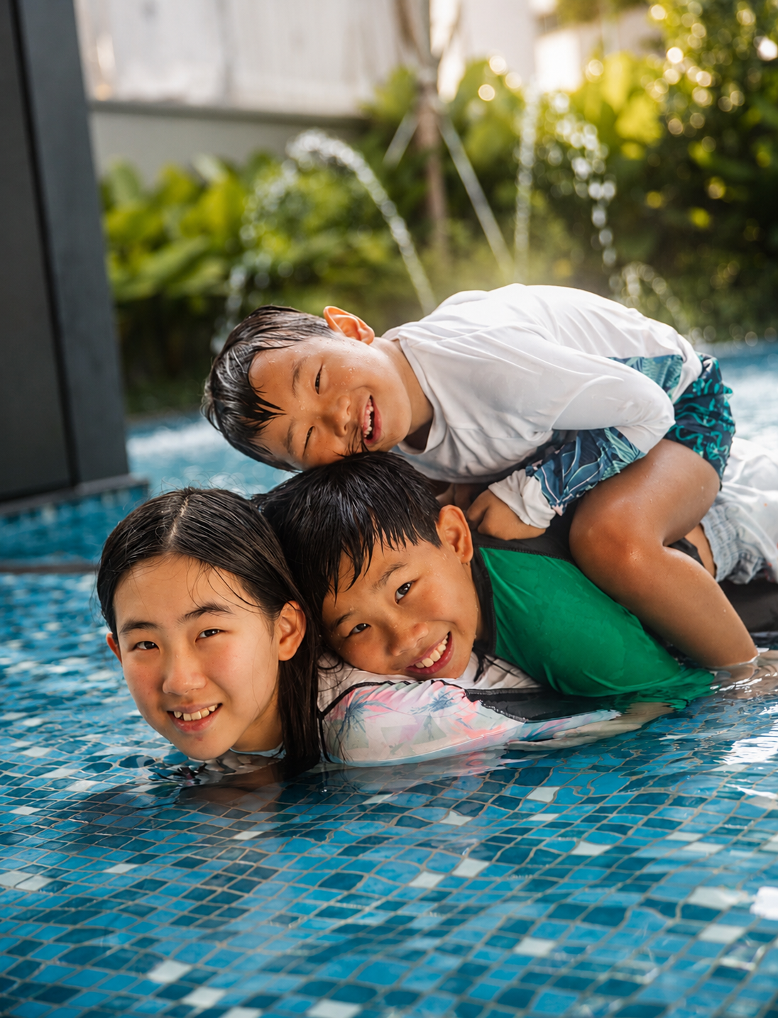 Three laughing Asian children playfully stacked on each other in a sunny outdoor pool.