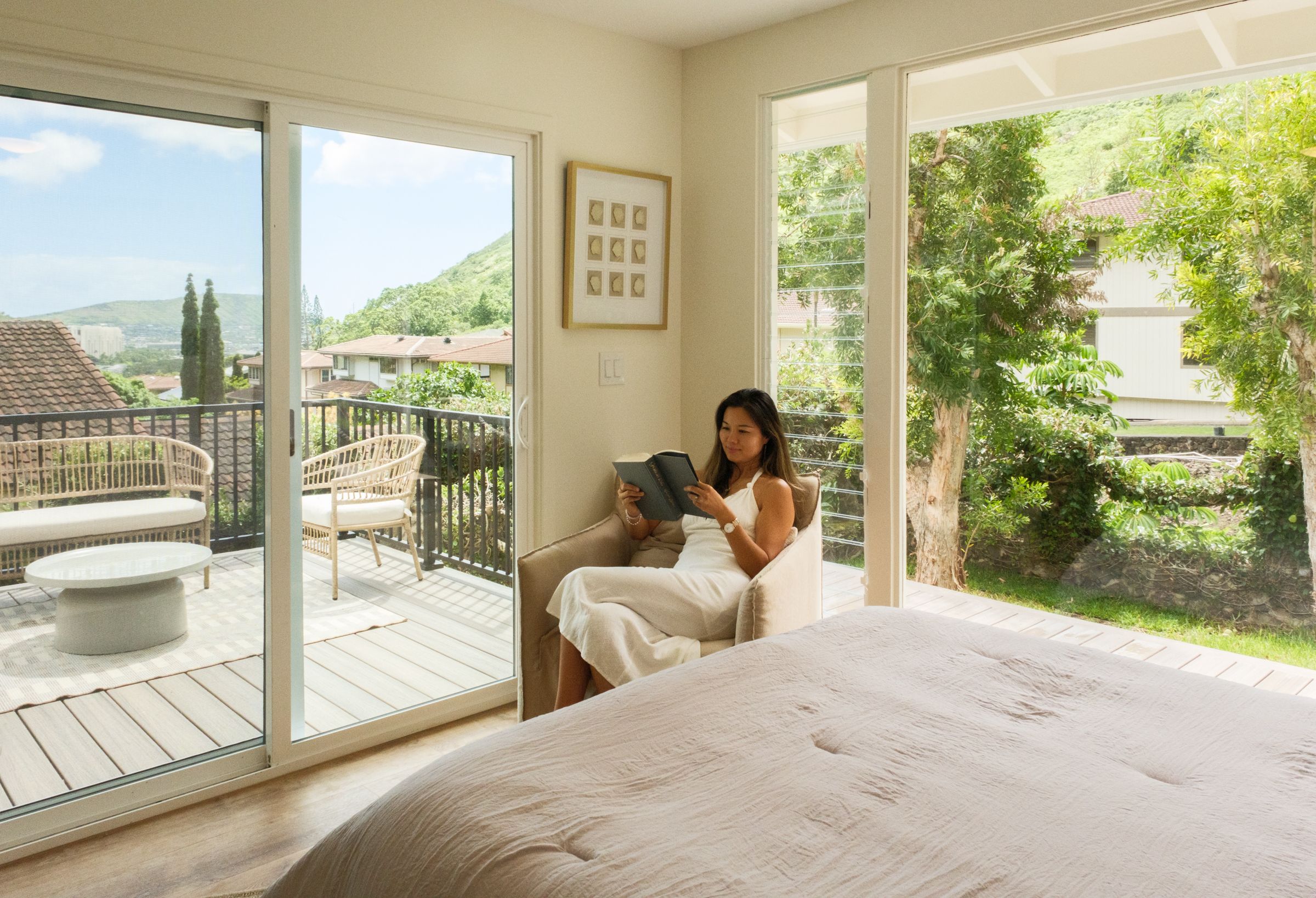 Sunlit bedroom with a woman seated beside sliding glass doors overlooking a terrace and garden.
