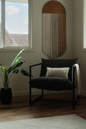 Dark accent chair with a cushion beneath a rounded wall mirror, beside a window and leafy plant.