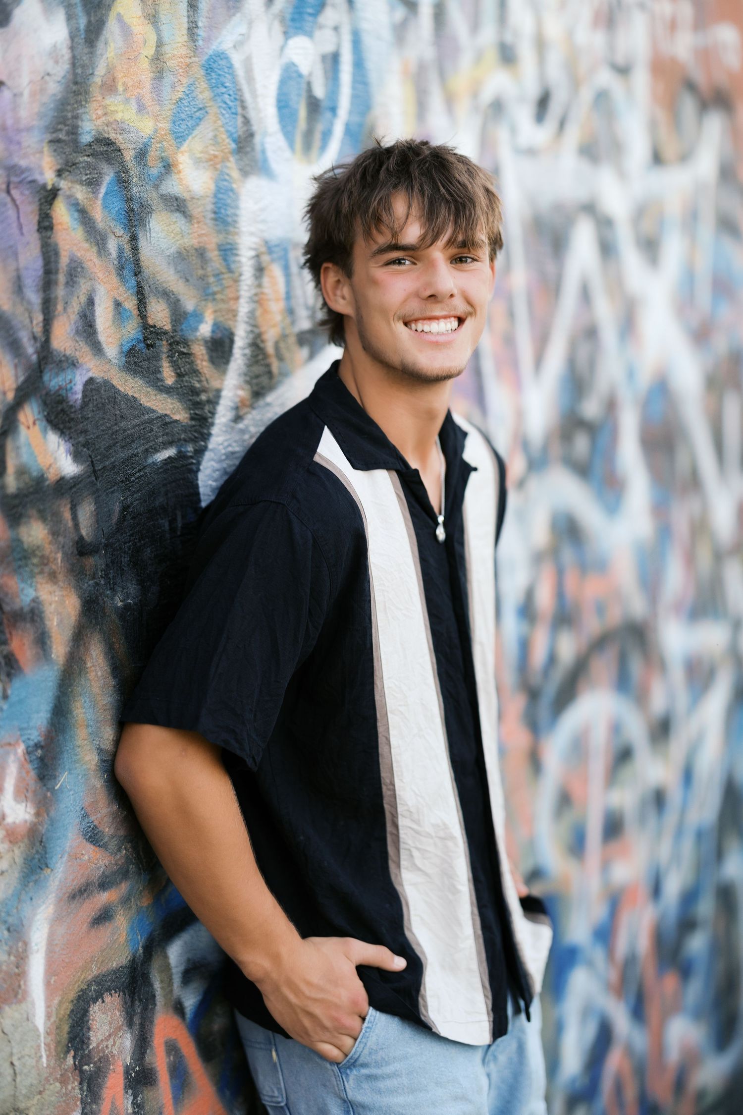 Smiling young man in black shirt leaning against colorful graffiti wall with hands in pockets.