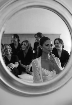 Black and white mirror reflection of a bride smiling during wedding preparations, surrounded by bridesmaids in dark robes.
