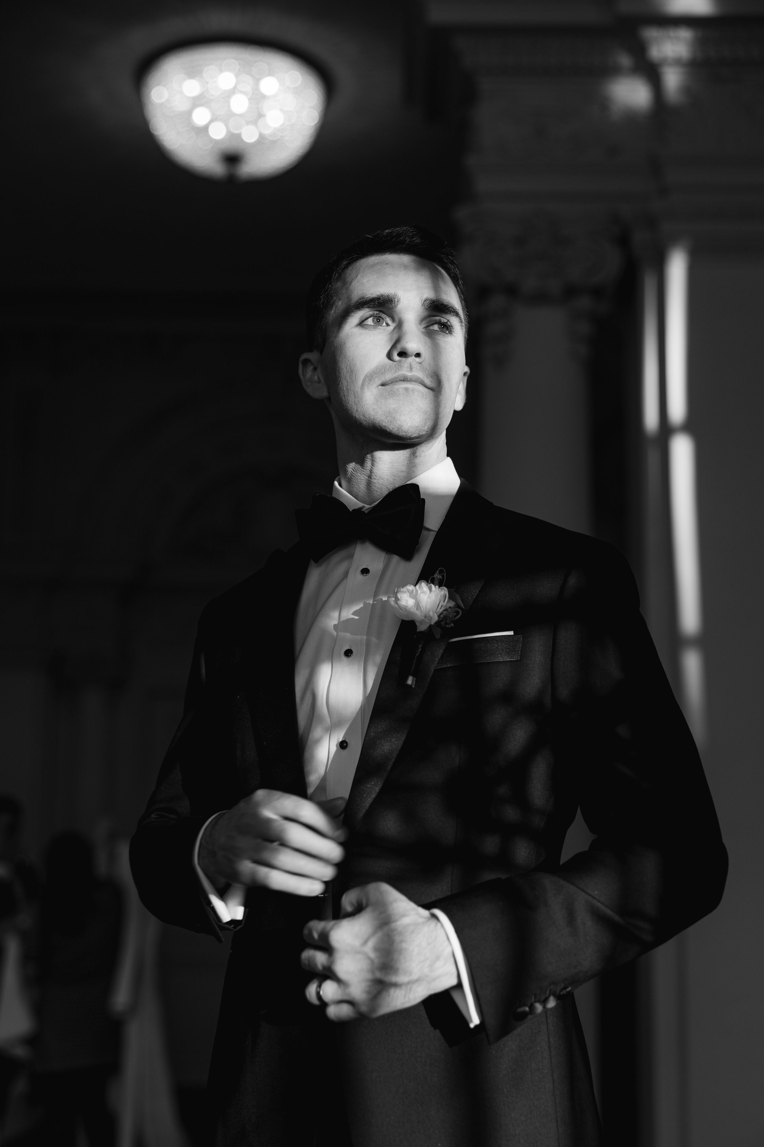 Black and white portrait of a groom in a tuxedo with bow tie and boutonniere, adjusting his jacket indoors.