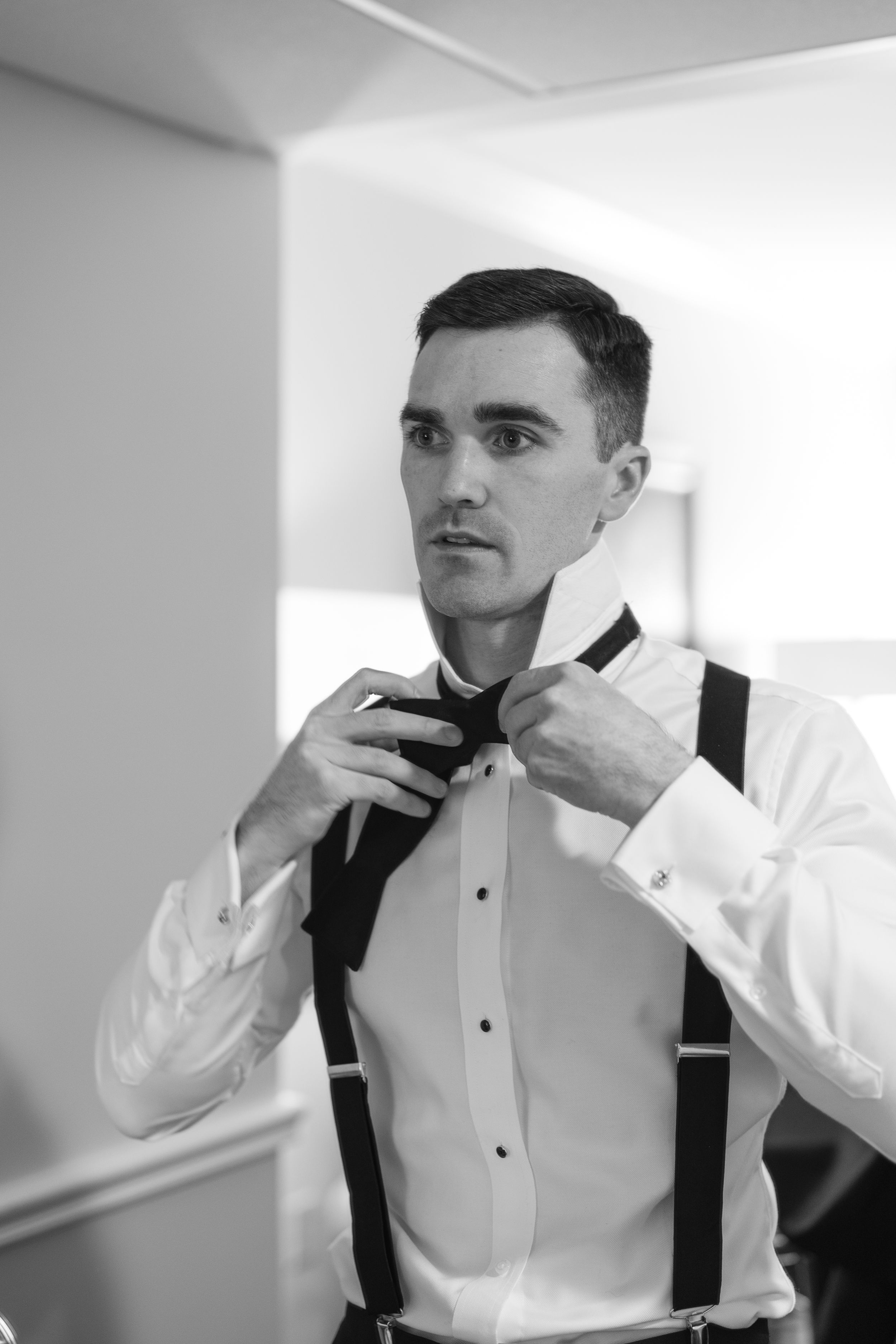Black and white photo of a groom adjusting his bow tie wearing suspenders and a white dress shirt.