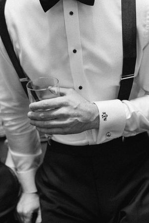 Black and white photo of groom in tuxedo shirt with suspenders and bow tie, holding a glass with monogrammed cufflinks.