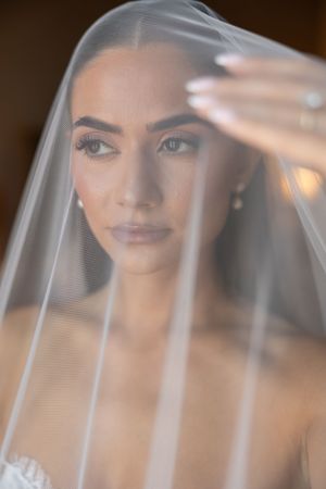 Bride in white lace gown holds sheer veil over face, showcasing elegant makeup and diamond ring.