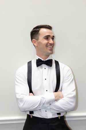 Smiling young man in white tuxedo shirt, black bow tie and suspenders, arms crossed, posing against a light wall.