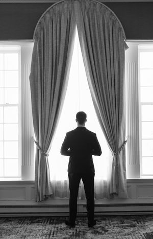 Black and white photo of a man in a suit standing before elegant draped curtains gazing out a large arched window.