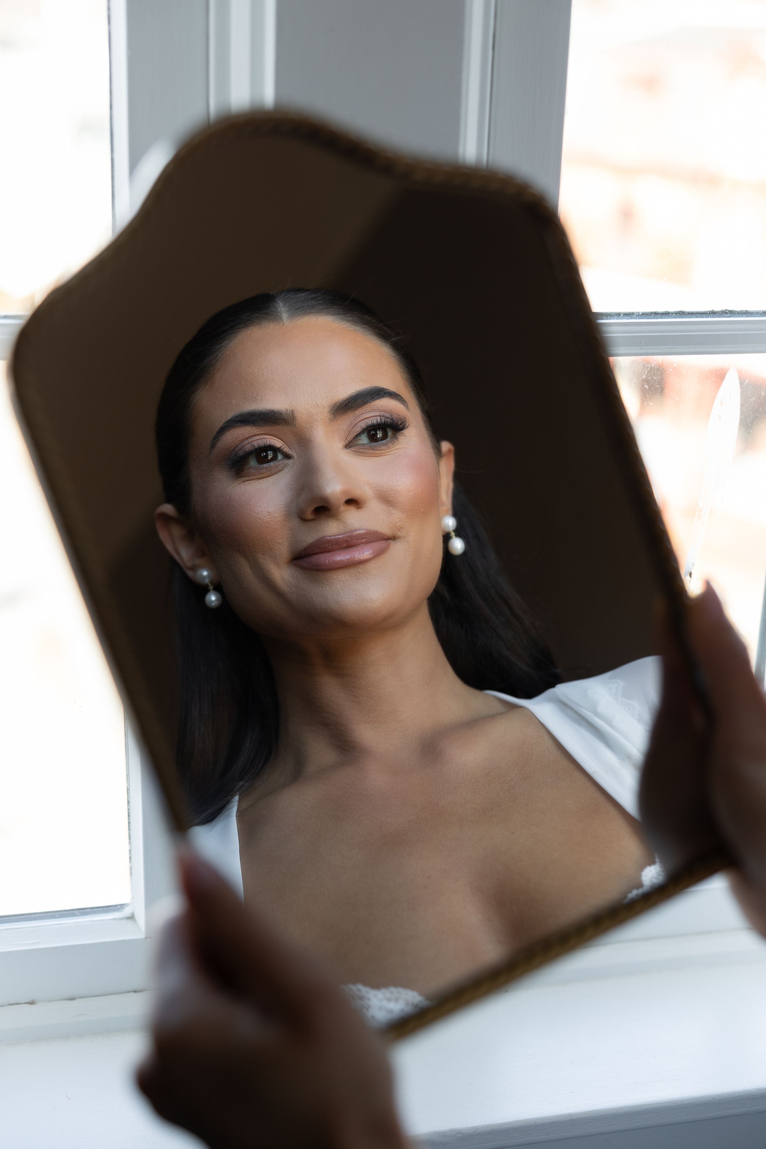 A bride with pearl earrings and elegant makeup gazes at her reflection in a vintage handheld mirror near a window.