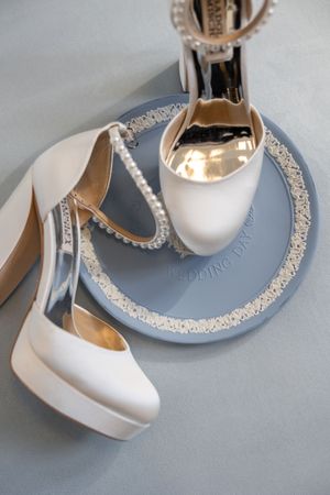 Elegant white satin bridal heels with pearl ankle straps displayed on a decorative wedding day plate.