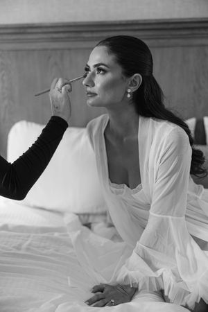Black and white photo of a bride in white robe getting makeup applied while sitting on a bed.