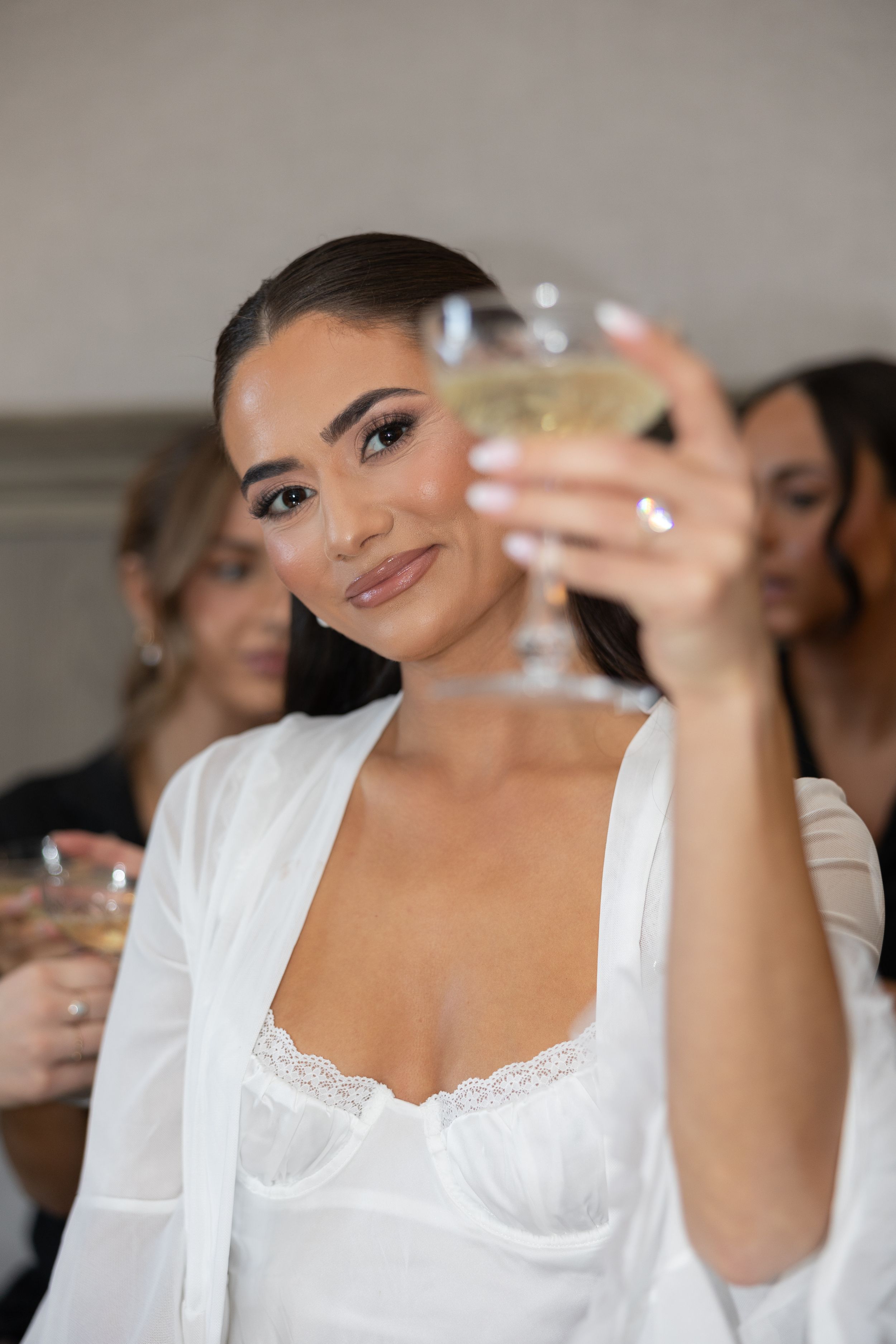 Bride in white lace robe raising champagne glass with bridesmaids during wedding morning preparations.