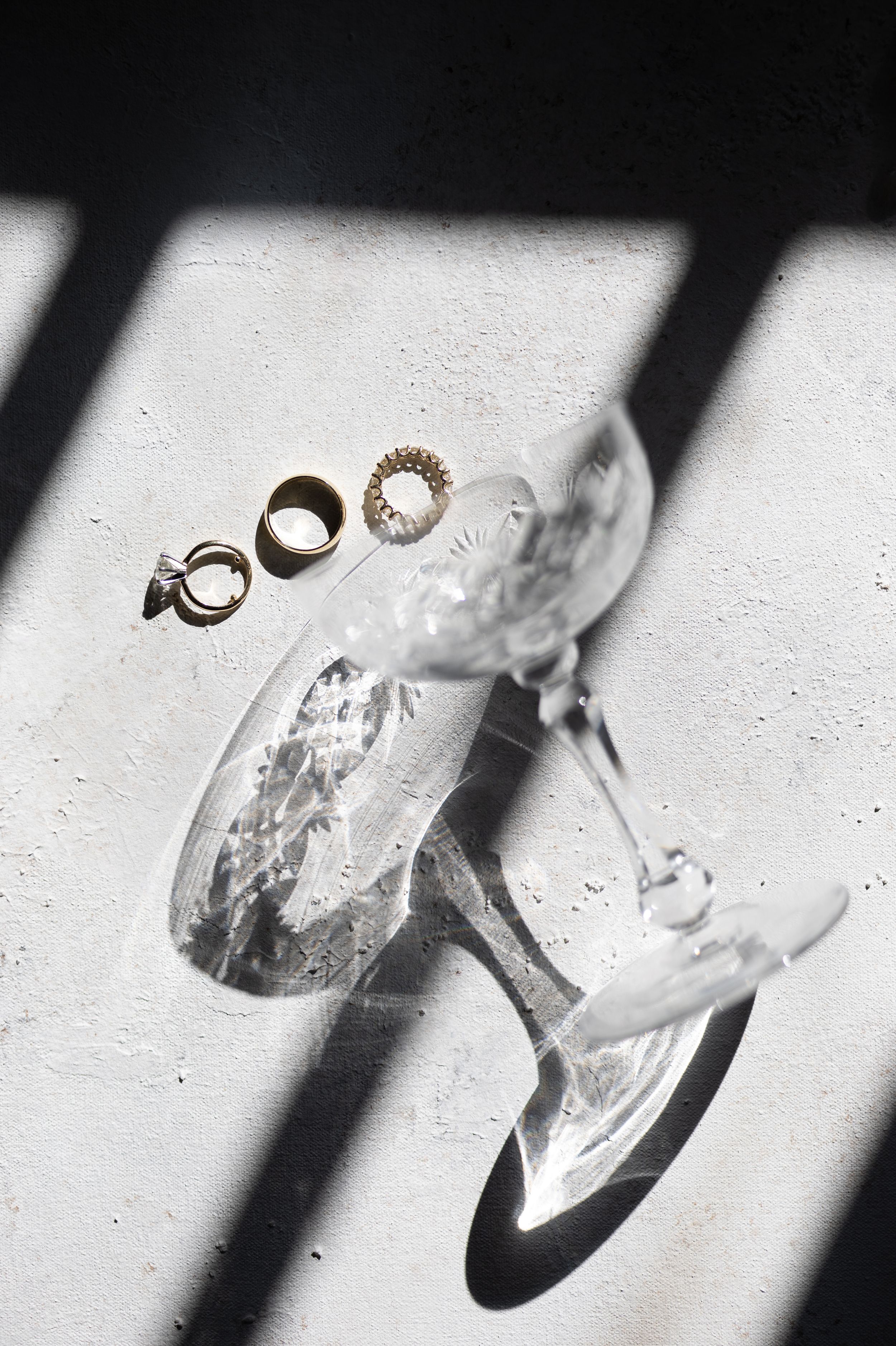 Crystal glass vase with gold wedding rings displayed on white surface with dramatic shadow patterns from window light.