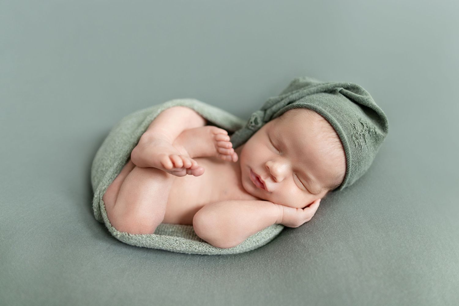 Sleeping newborn baby in sage green wrap and hat posed on matching green backdrop in studio photography.