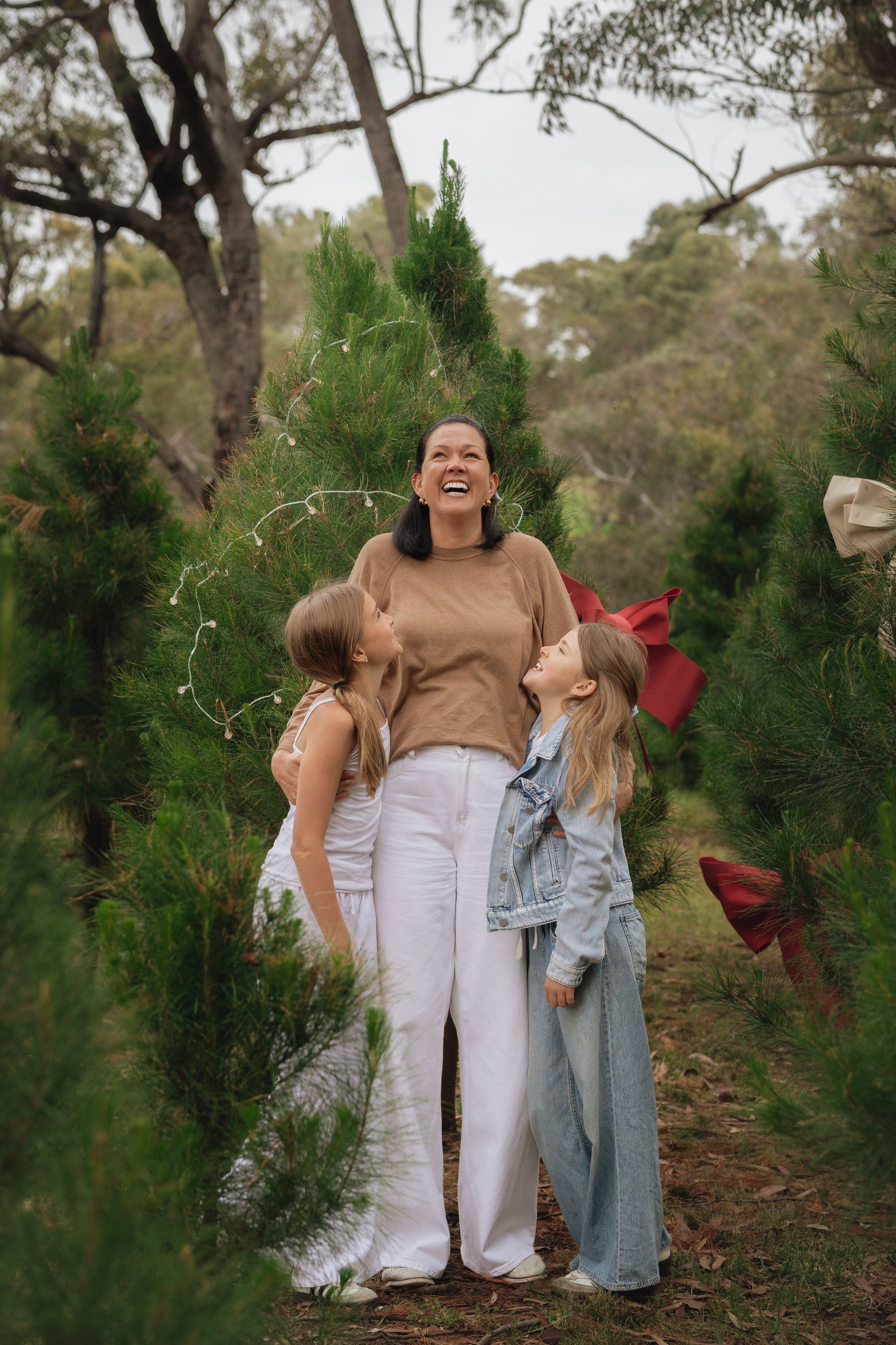 A laughing woman in a beige sweater and white pants stands with two girls amongst Christmas trees in Kenthurst during Christmas mini photos.