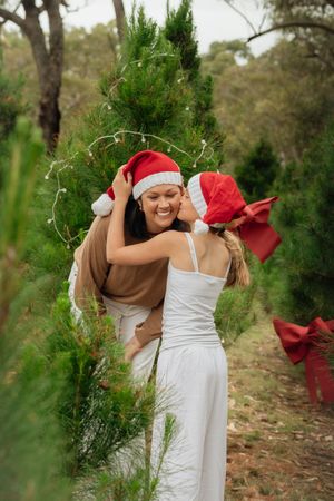 A girl and her mother in Santa hats embrace joyfully among green Christmas trees in Kenthurst, one wearing white dress, other in brown top.