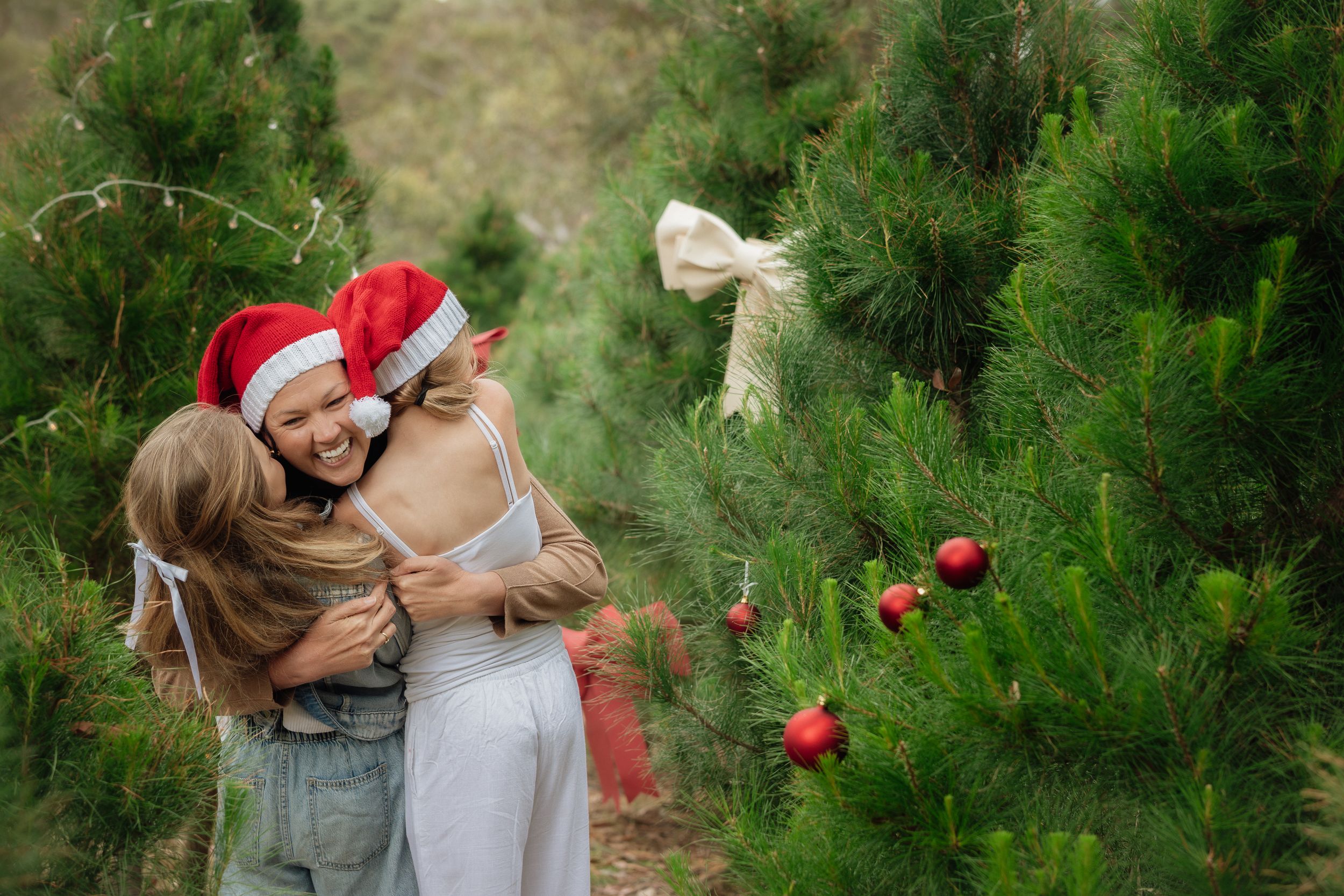 Two girls in Santa hats joyfully hug a woman among decorated Christmas trees at a festive tree farm during Christmas mini photos at Tomalong