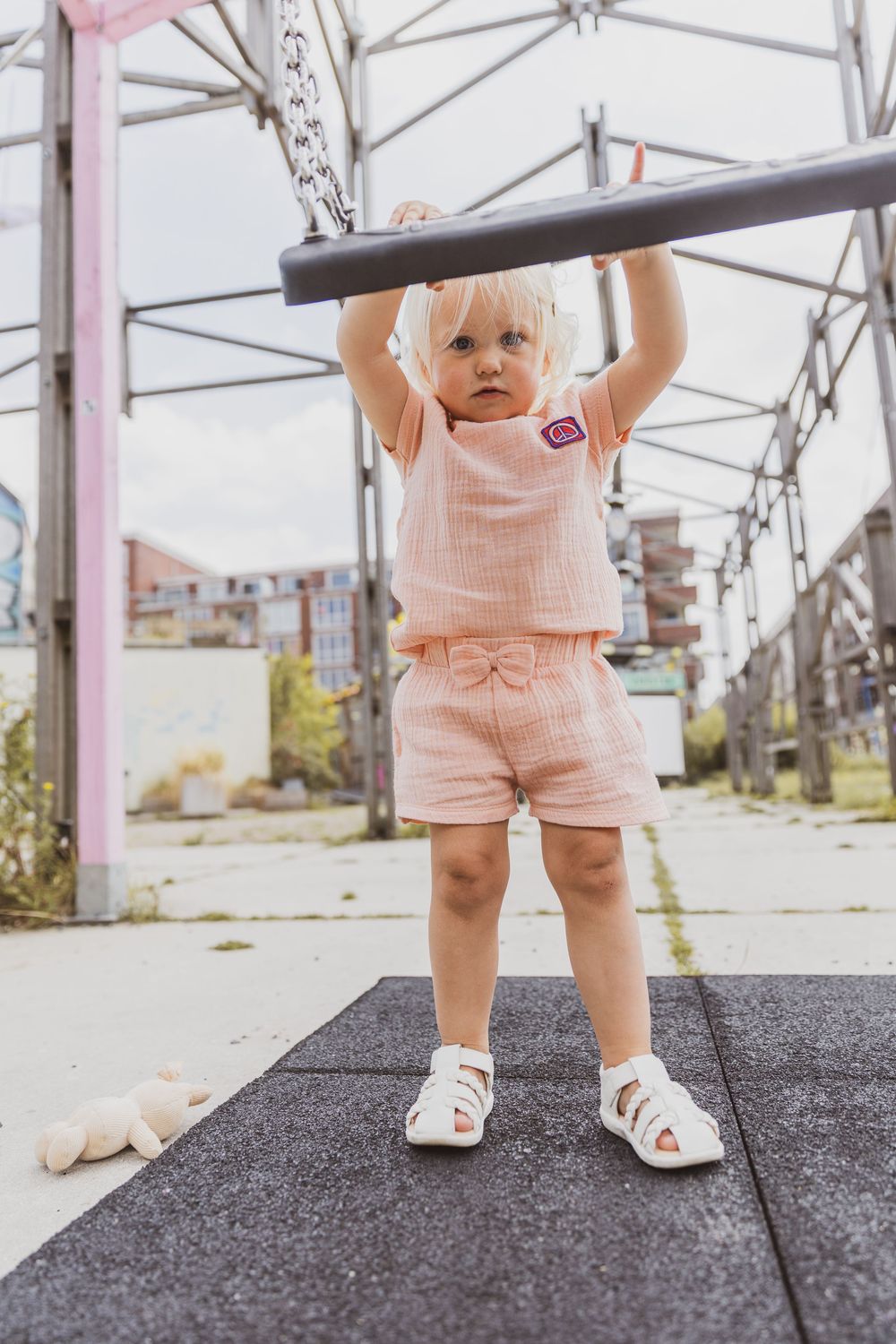 Toddler girl in pink outfit holding playground bar, standing on rubber mat outdoors.