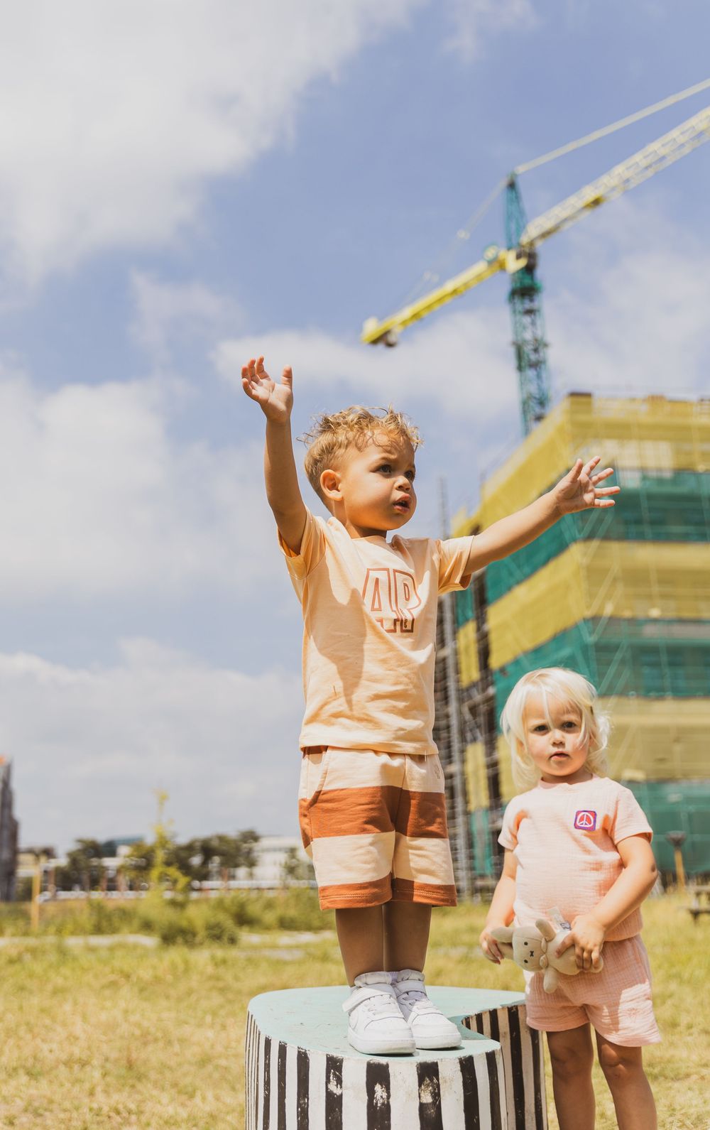 Two toddlers outdoors near a construction site, one standing on a striped box with arms raised, wearing matching orange outfits.