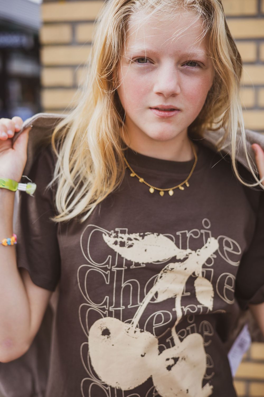 Young girl with blonde hair wearing a brown cherry graphic t-shirt and gold necklace, standing outdoors near a brick wall.