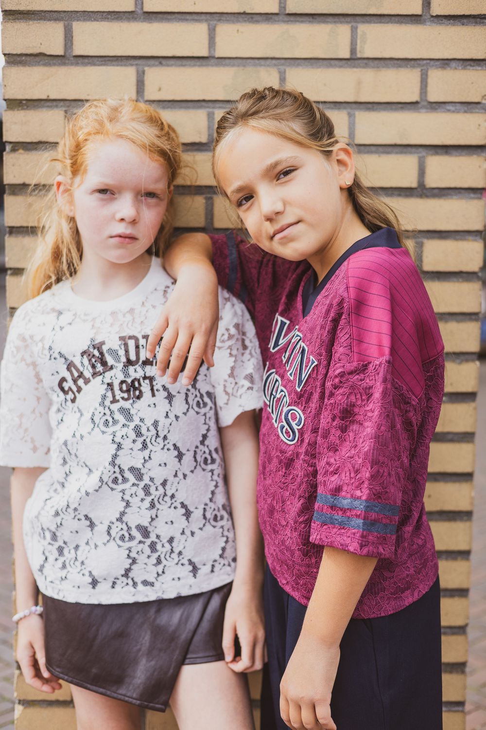 Two young girls posing against a brick wall, one in white lace top and one in purple jersey, smiling at camera.