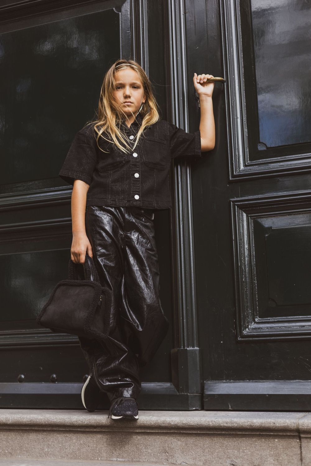 Young girl in black button-up shirt and shiny black pants poses confidently by a dark door holding a small purse.