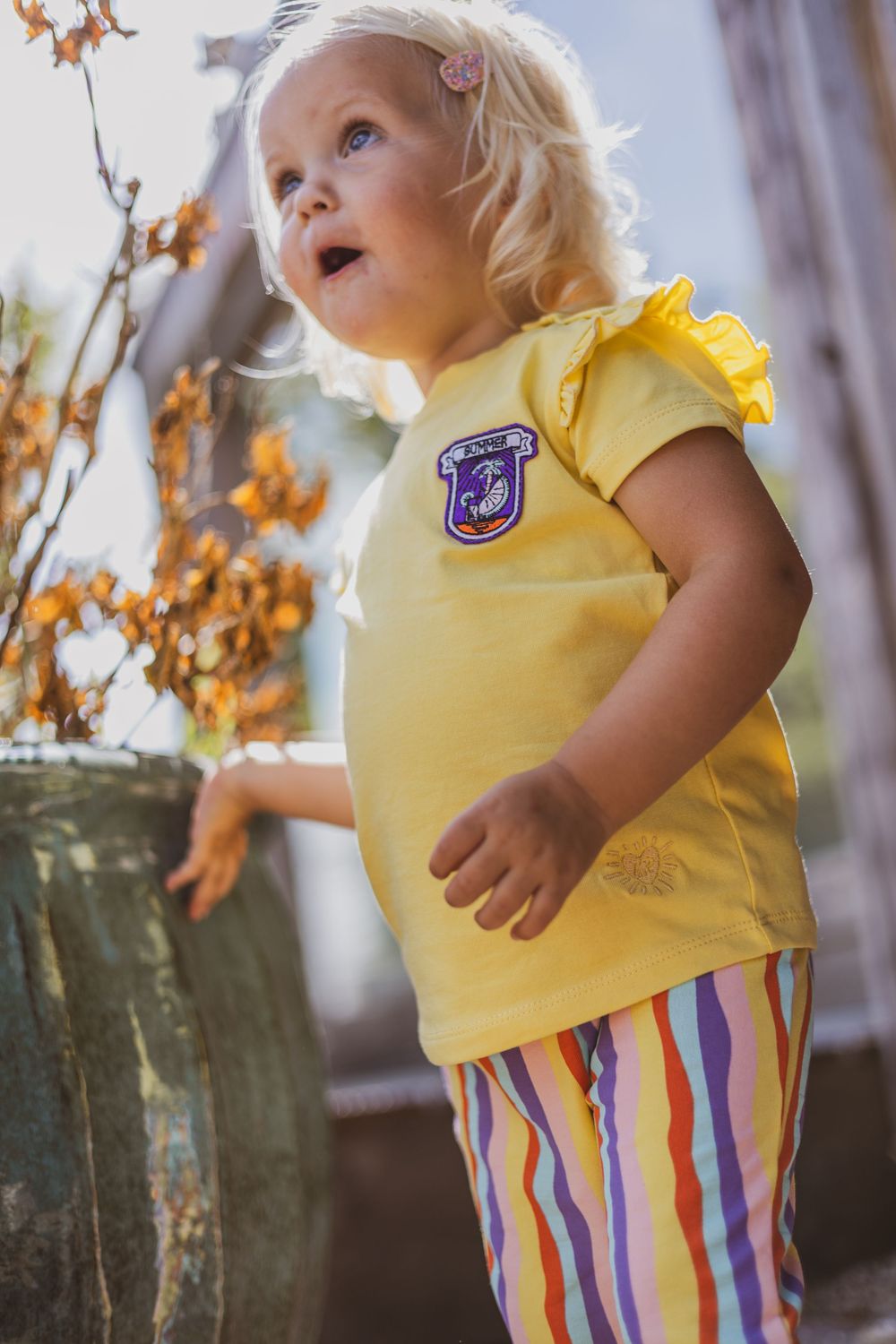 Toddler girl in yellow top and striped pants standing outdoors near autumn plants in natural sunlight.