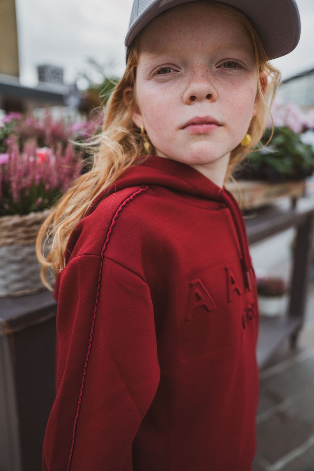 Red-haired girl in red hoodie and gray cap standing near flower display, looking over shoulder.