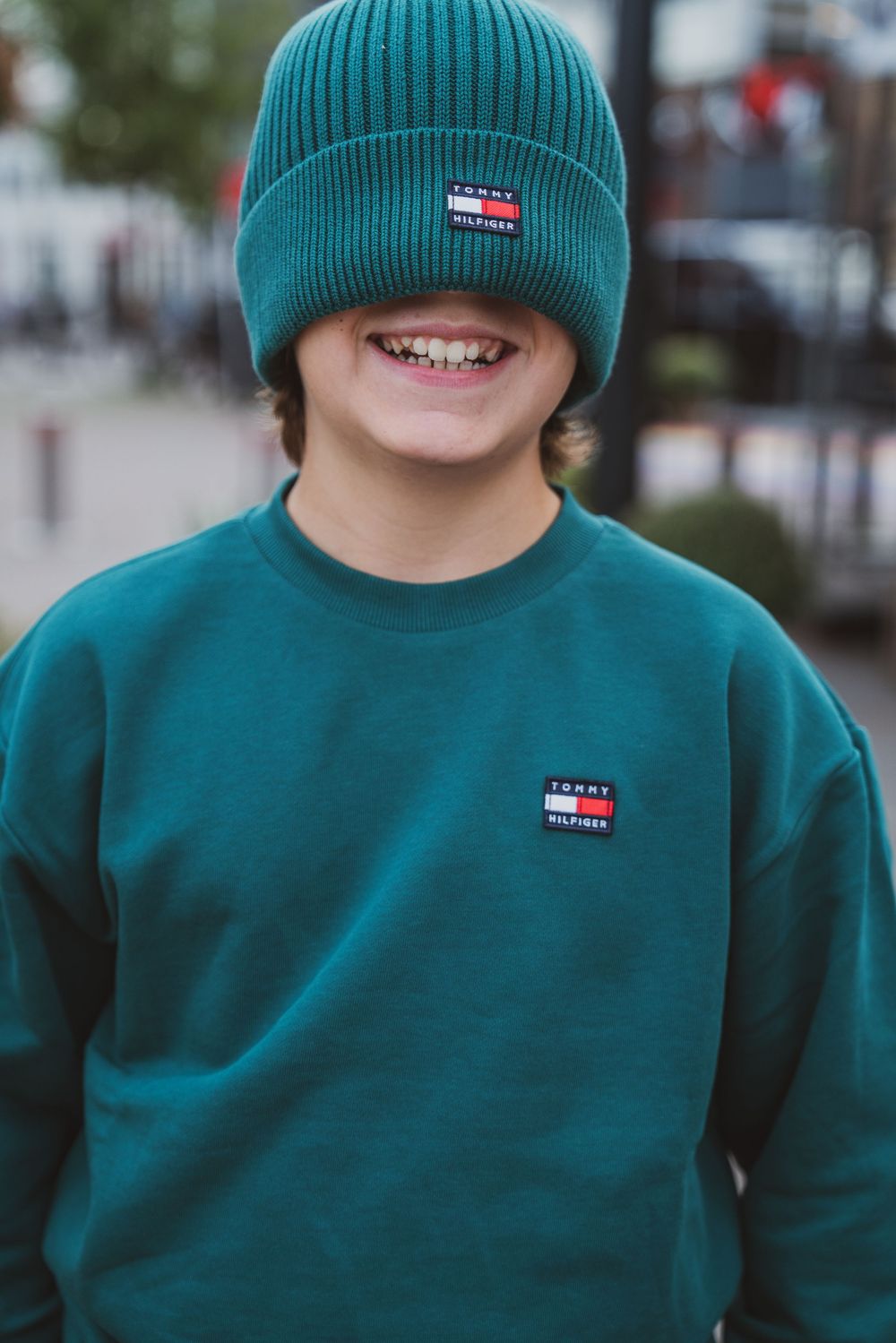 Smiling boy wearing a teal Tommy Hilfiger beanie and matching sweatshirt outdoors.