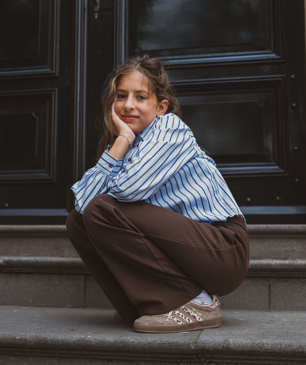 Young girl in blue striped shirt and brown pants squatting on stone steps, smiling with hand on chin.
