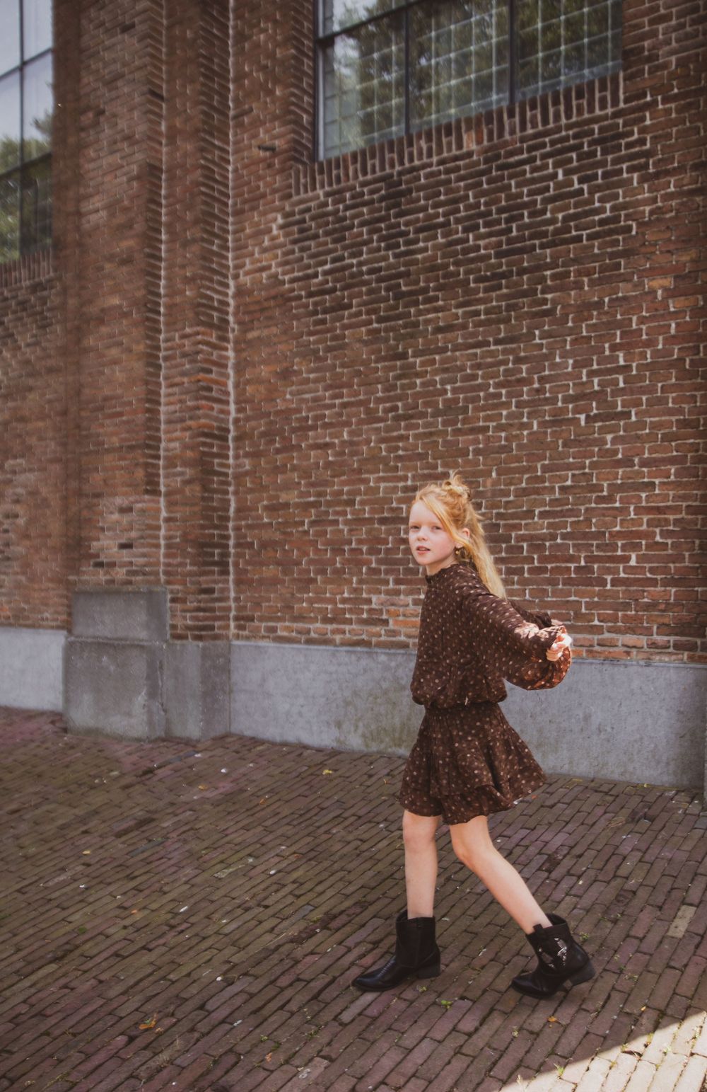 A young girl in a brown floral dress and black boots walks along a brick wall outdoors.