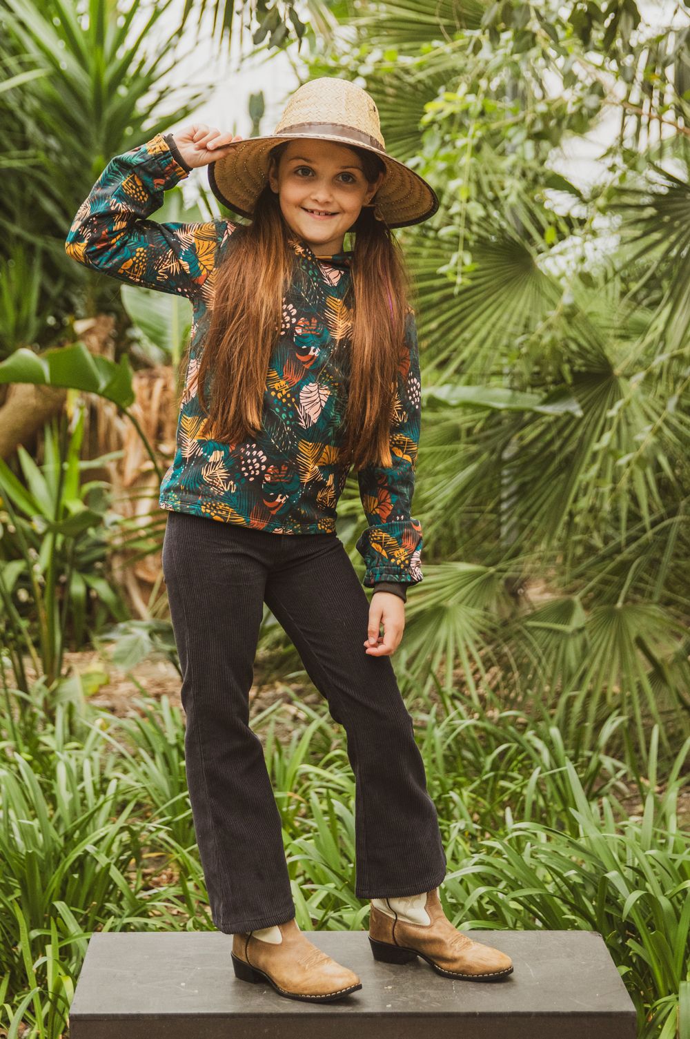 Young girl in floral top, dark jeans, tan boots and straw hat posing outdoors among tropical green plants.