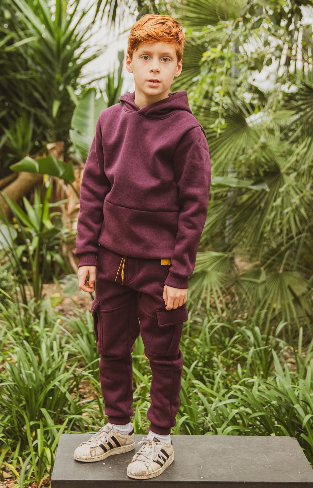 Boy wearing a burgundy hoodie and cargo joggers set with white Adidas sneakers, standing outdoors among tropical plants.