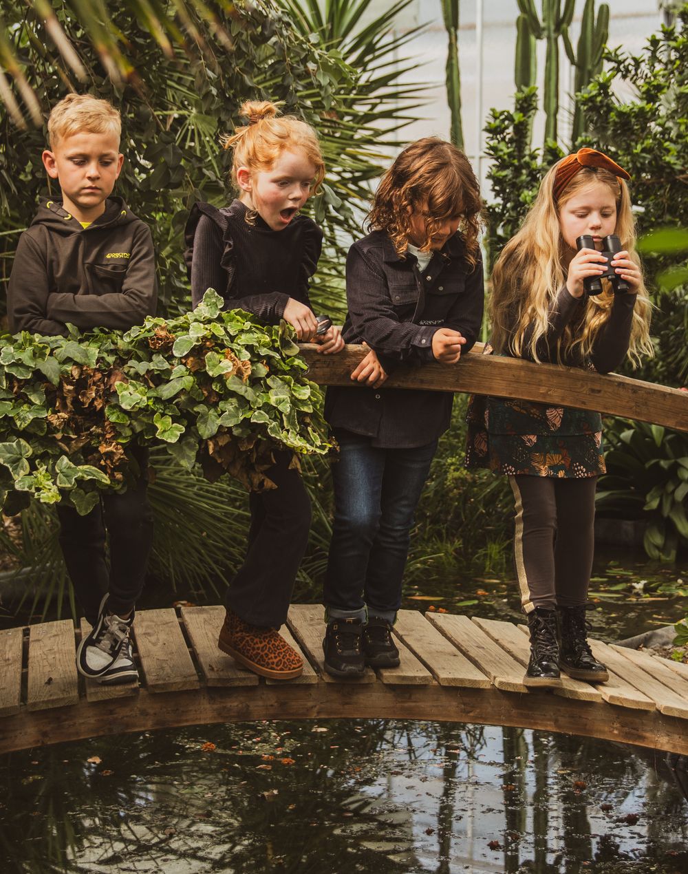 Four children explore a lush greenhouse garden, standing on a wooden bridge over a pond surrounded by tropical plants.