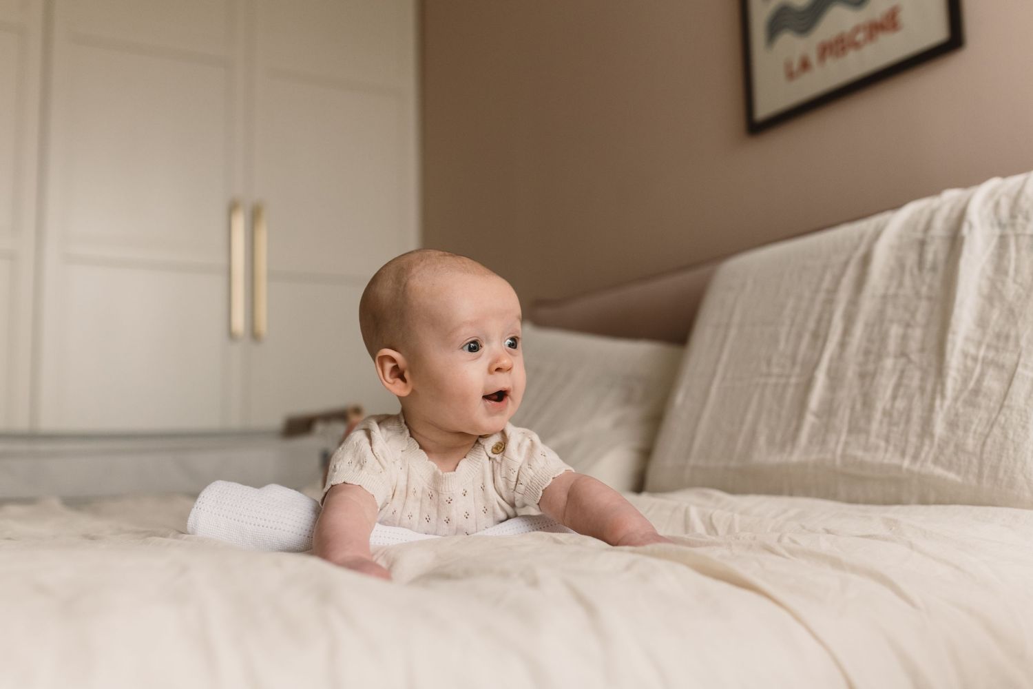 A curious baby in a white onesie doing tummy time on a cream bed with soft white bedding.